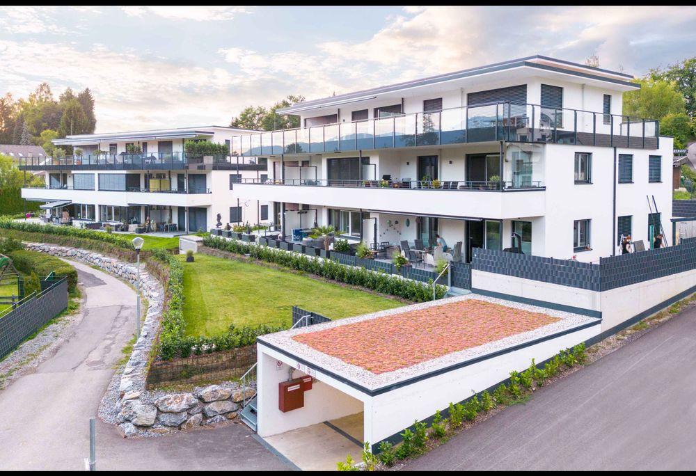 Willkommen in unserem modern und elegant gestalteten Wohnanlage, die von uns als Bauträger errichtet und verkauft wurden. Dieses Bild zeigt das schöne Äußere der modernen Gebäude, umgeben von Grün und Bäumen. Im Hintergrund können wir den klaren blauen Himmel und eine ferne Stadtlandschaft sehen. Diese Wohnanlage bietet luxuriöse Wohnräume, hochmoderne Annehmlichkeiten und eine friedliche Umgebung, die die Bewohner ihr Zuhause nennen können.