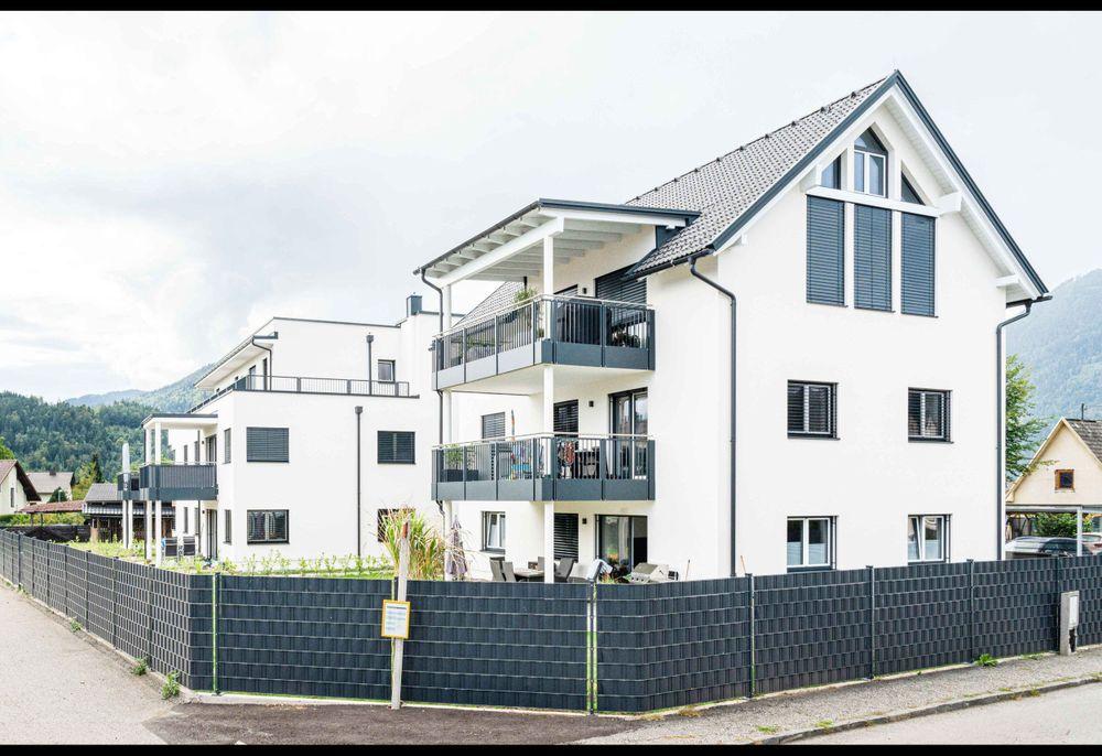 Willkommen in unserem modern und elegant gestalteten Wohnanlage, die von uns als Bauträger errichtet und verkauft wurden. Dieses Bild zeigt das schöne Äußere der modernen Gebäude, umgeben von Grün und Bäumen. Im Hintergrund können wir den klaren blauen Himmel und eine ferne Stadtlandschaft sehen. Diese Wohnanlage bietet luxuriöse Wohnräume, hochmoderne Annehmlichkeiten und eine friedliche Umgebung, die die Bewohner ihr Zuhause nennen können.