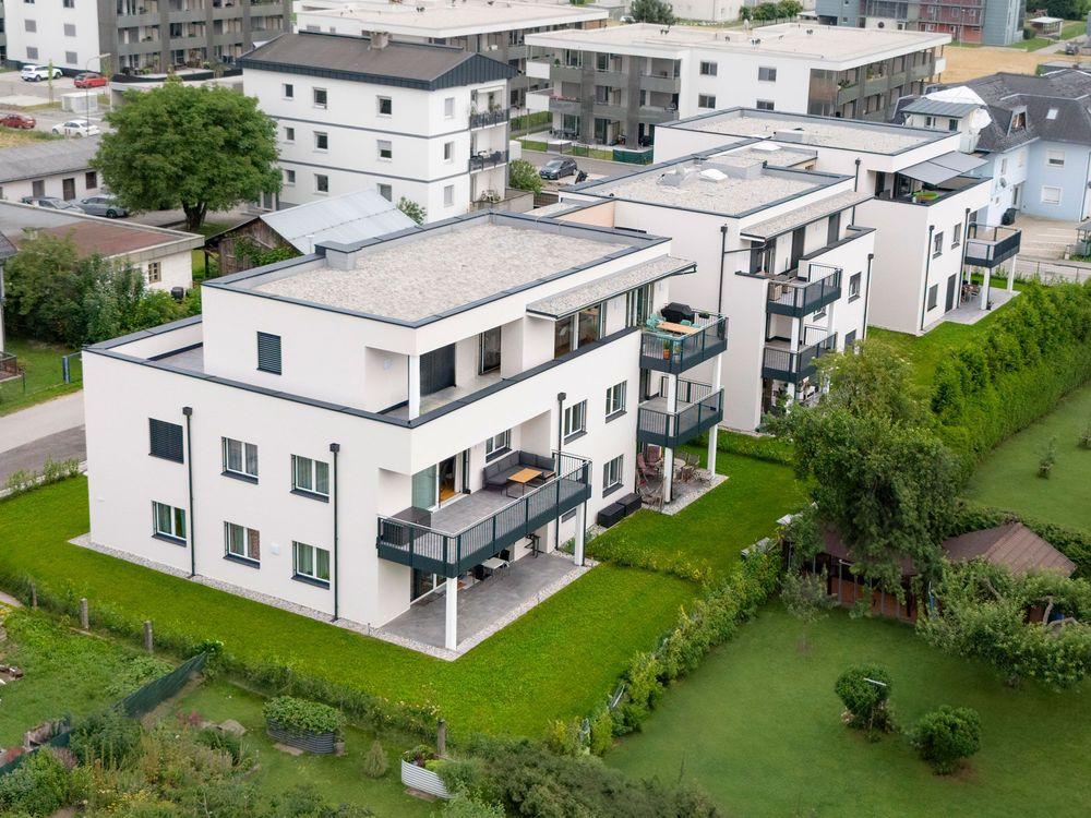 Willkommen in unserem modern und elegant gestalteten Wohnanlage, die von uns als Bauträger errichtet und verkauft wurden. Dieses Bild zeigt das schöne Äußere der modernen Gebäude, umgeben von Grün und Bäumen. Im Hintergrund können wir den klaren blauen Himmel und eine ferne Stadtlandschaft sehen. Diese Wohnanlage bietet luxuriöse Wohnräume, hochmoderne Annehmlichkeiten und eine friedliche Umgebung, die die Bewohner ihr Zuhause nennen können.