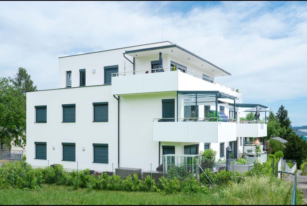 Willkommen in unserem modern und elegant gestalteten Wohnanlage, die von uns als Bauträger errichtet und verkauft wurden. Dieses Bild zeigt das schöne Äußere der modernen Gebäude, umgeben von Grün und Bäumen. Im Hintergrund können wir den klaren blauen Himmel und eine ferne Stadtlandschaft sehen. Diese Wohnanlage bietet luxuriöse Wohnräume, hochmoderne Annehmlichkeiten und eine friedliche Umgebung, die die Bewohner ihr Zuhause nennen können.